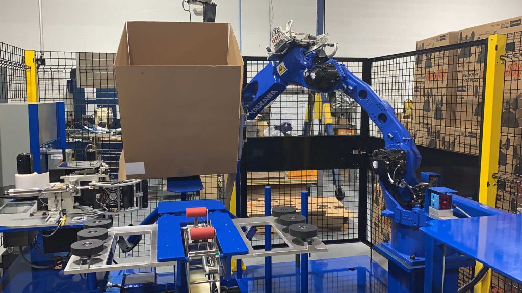 Robotic case erector in action, lifting a cardboard box with a blue Yaskawa robotic arm, demonstrating flexibility and efficiency in handling various case sizes.