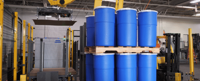 Robotic industrial palletizer system stacking blue plastic drums on a wooden pallet.